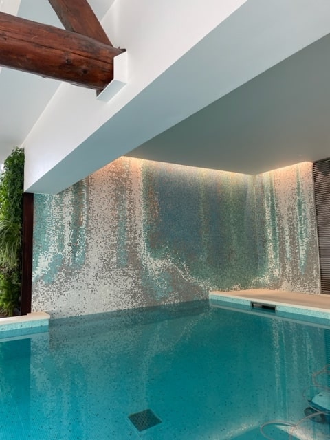 Piscine intérieure moderne avec un mur en mosaïque dégradé bleu et blanc, éclairage indirect et poutre en bois rustique.